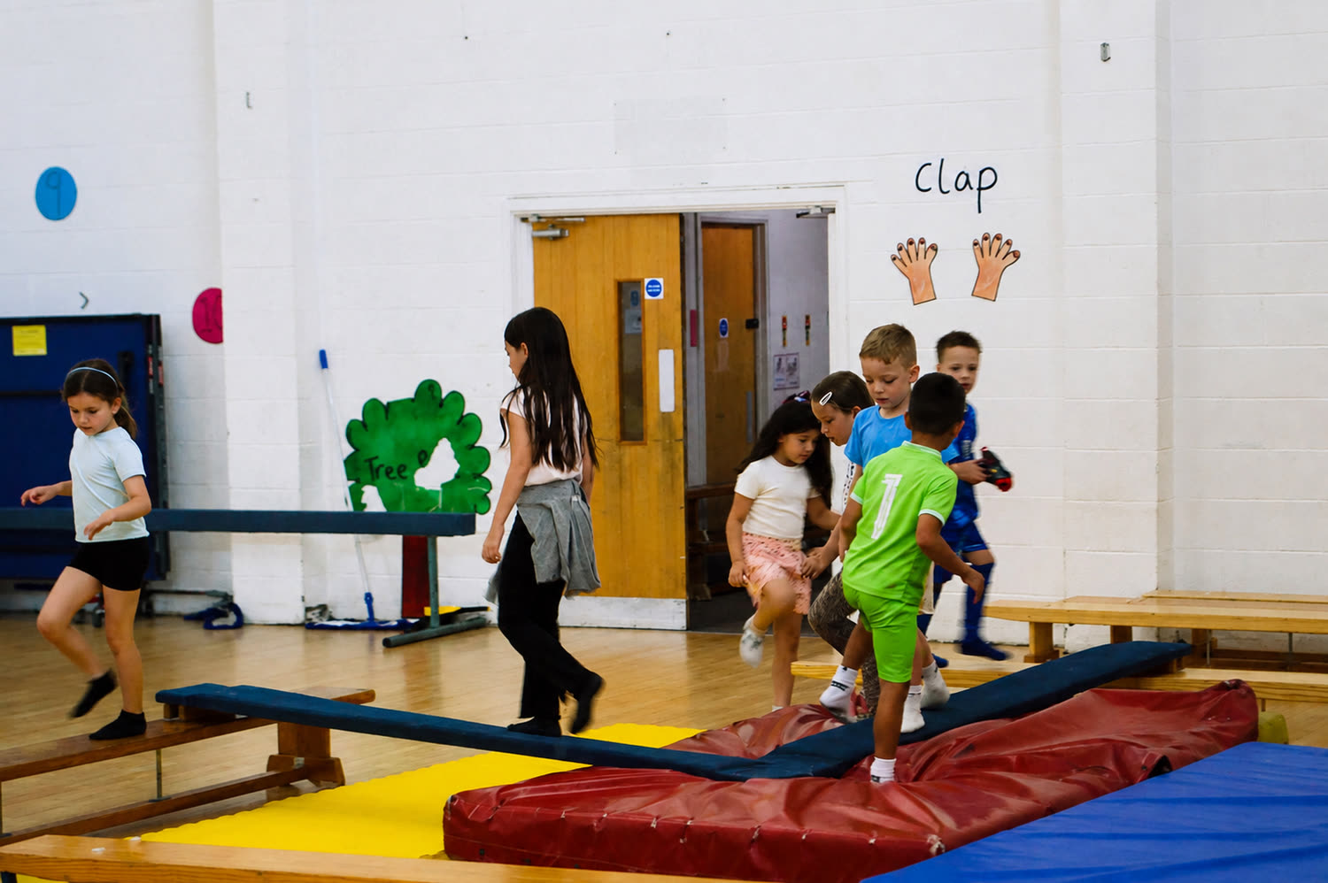 A group of children crossing the balance beams and crash mats in the hall